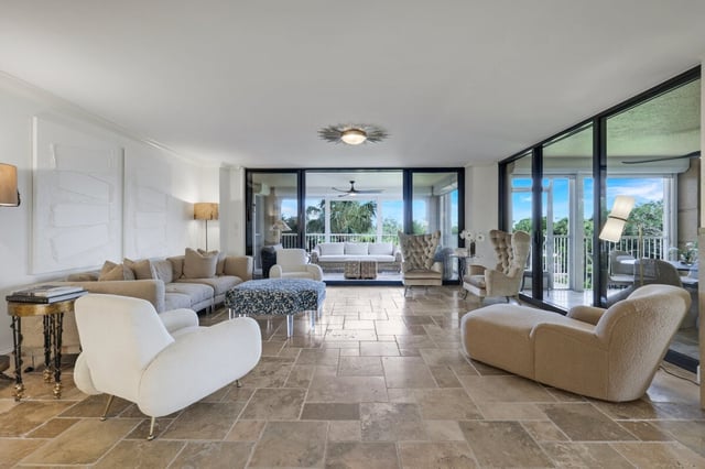 Spacious living room with travertine floors, neutral sofas and armchairs, and floor-to-ceiling windows opening to a green view