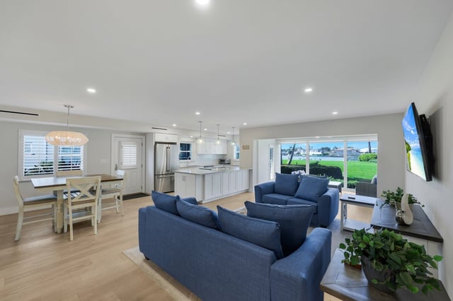 Open-plan living room with navy sofas, white kitchen and dining table, and sliding doors to a waterfront yard