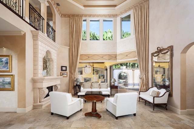 Two-story living room with carved stone fireplace, white upholstered armchairs and tall draped windows over travertine floors