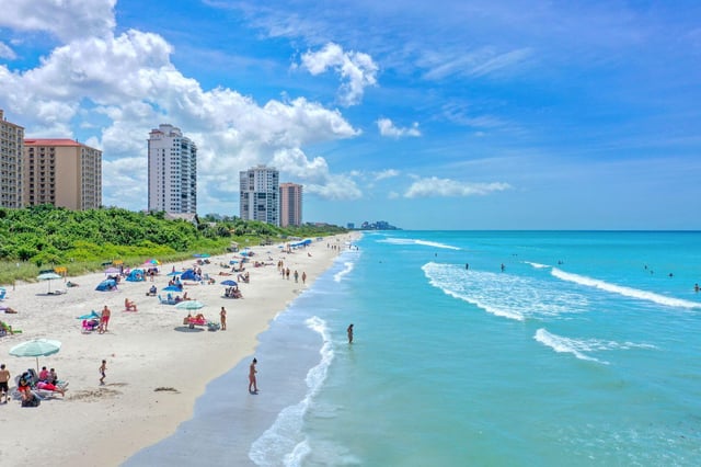 Aerial view of a wide white sand beach with beachgoers, turquoise Gulf surf and high-rise condos in Naples