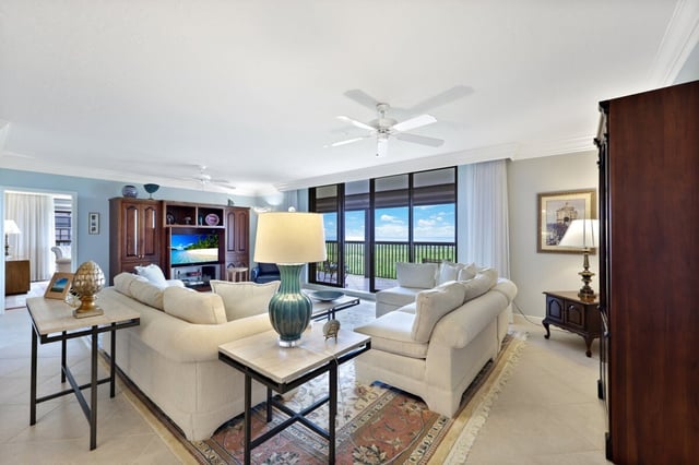 Bright condo living room with cream sofas, ceiling fan and floor-to-ceiling windows framing a Gulf view
