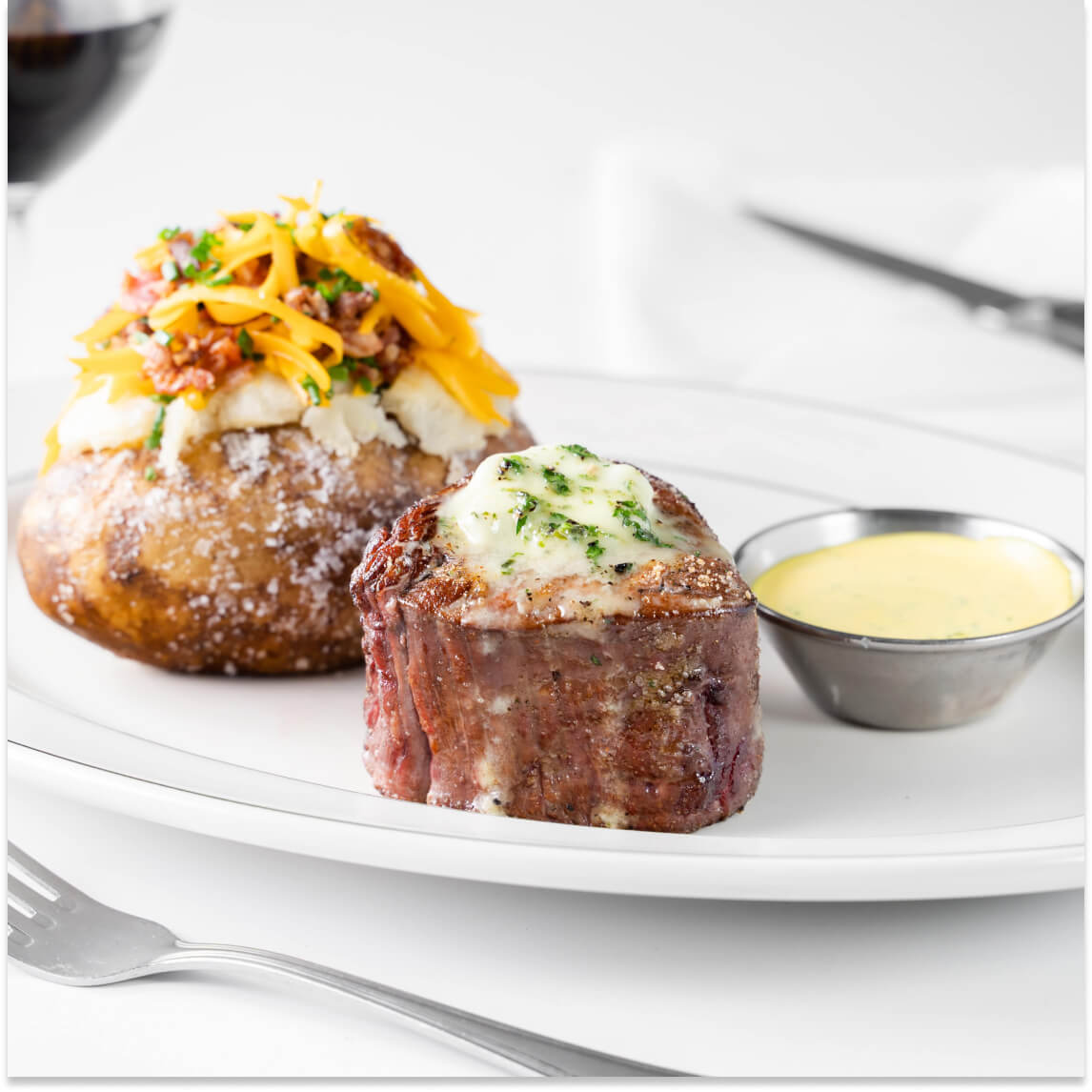 Filet topped with herb butter beside a loaded baked potato and a cup of yellow sauce on a white plate