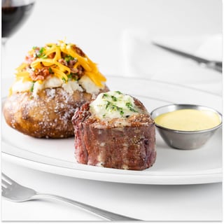 Filet topped with herb butter beside a loaded baked potato and a cup of yellow sauce on a white plate
