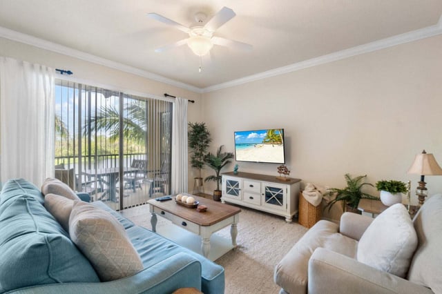 Bright coastal living room with teal sofa, ceiling fan, wall-mounted TV and sliding doors to a palm-shaded balcony