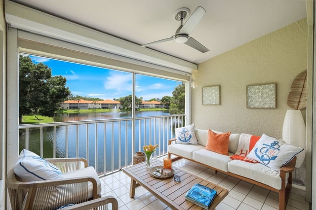 Screened lanai with white sofa, ceiling fan and coffee table overlooking a lake and tile-roofed condos
```