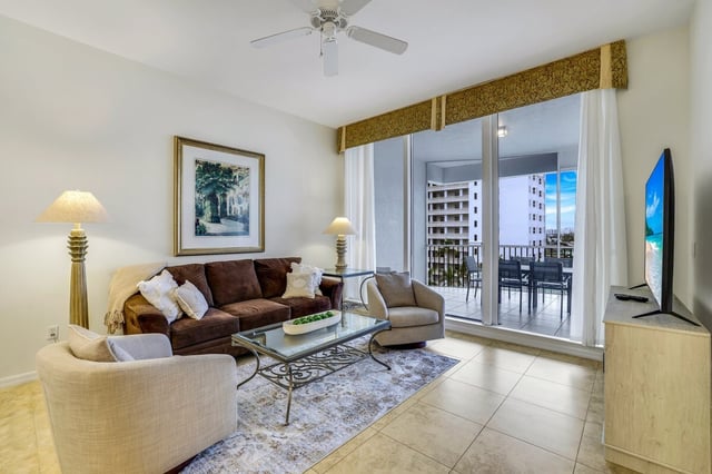 Condo living room with brown sofa, glass coffee table and ceiling fan opening to a balcony with resort views