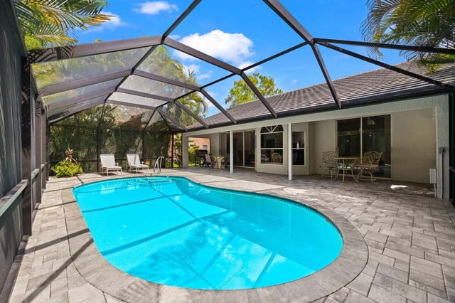 Screened lanai with a bright blue pool, paver deck, lounge chairs and patio dining beside a tile-roof home
