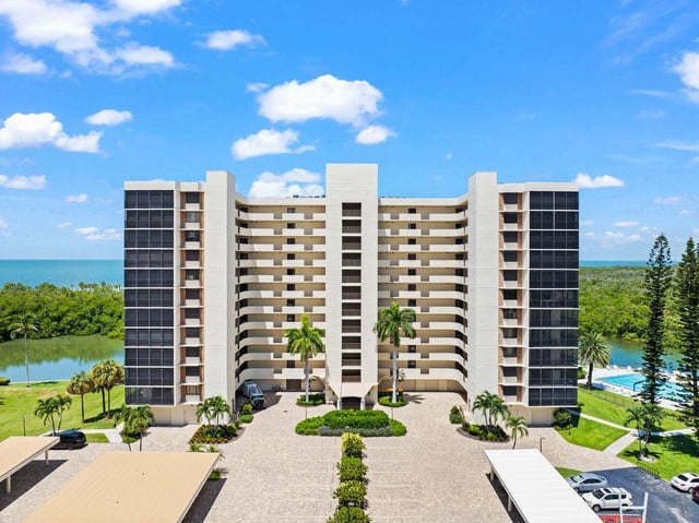 Aerial view of a tall beachfront condominium tower with palm-lined driveway, pool and Gulf waters beyond