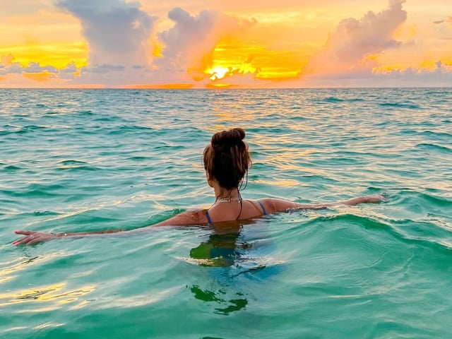 Woman floating with arms outstretched in turquoise Gulf water as a golden sunset lights the clouds
