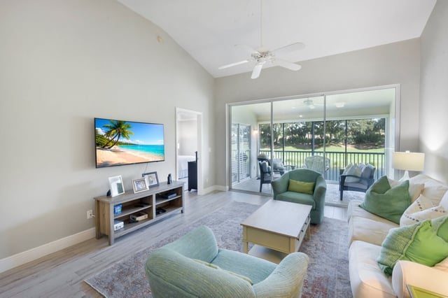 Bright vacation rental living room with vaulted ceiling, wall-mounted TV, green armchairs and sliding doors to a screened lanai overlooking a golf cou