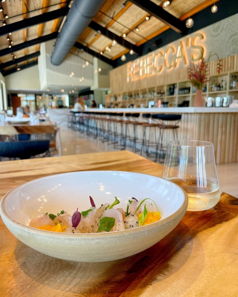A crudo dish in a ceramic bowl with white wine on a wood table inside Rebecca's airy bar dining room
