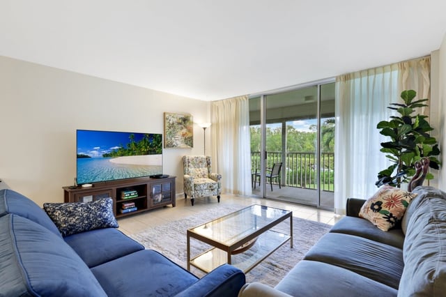 Bright condo living room with blue sectional sofas, glass coffee table and sliding doors to a green balcony view