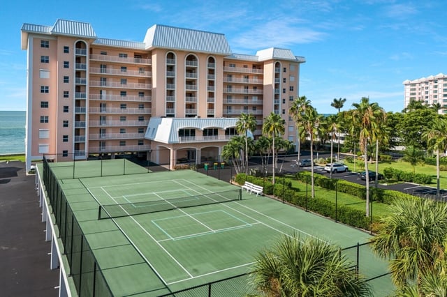 Green tennis courts beside a pink beachfront condominium tower lined with palms and the Gulf beyond