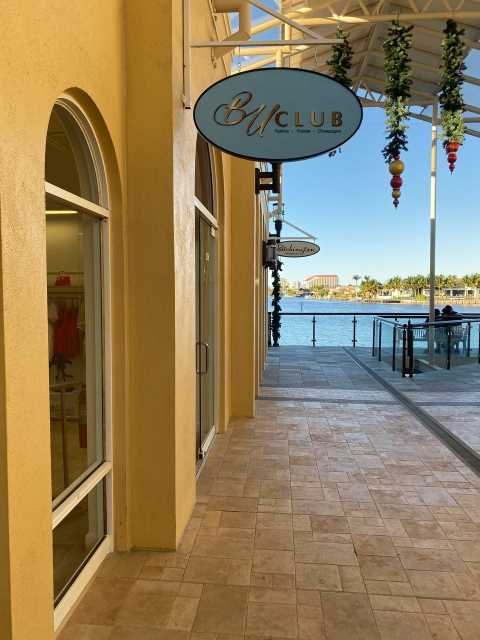 BU Club oval sign hanging above a yellow stucco waterfront shopping promenade with tile walkway and marina views