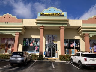 Pastel storefront of Alvin's Island Tropical Department Store with swimwear displays and cars parked out front under blue sky