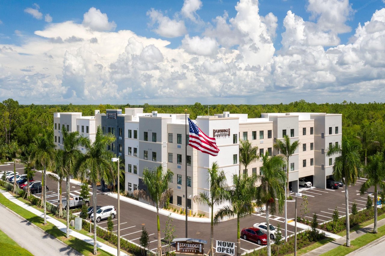 Aerial view of the white four-story Staybridge Suites Naples hotel with palm trees, an American flag and parking lot under bright clouds