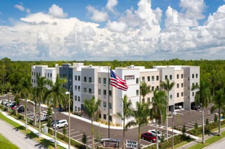 Aerial view of the white four-story Staybridge Suites Naples hotel with palm trees, an American flag and parking lot under bright clouds