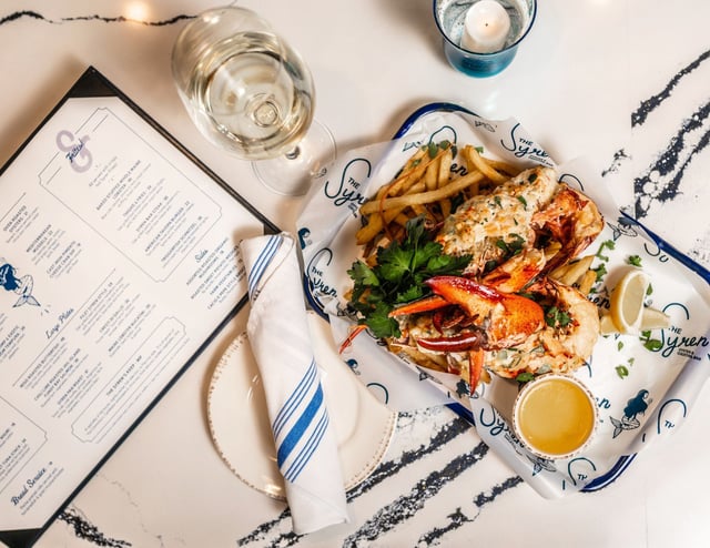 Overhead view of lobster with fries, lemon and drawn butter on branded paper beside white wine and a menu at The Syren Oyster & Cocktail Bar