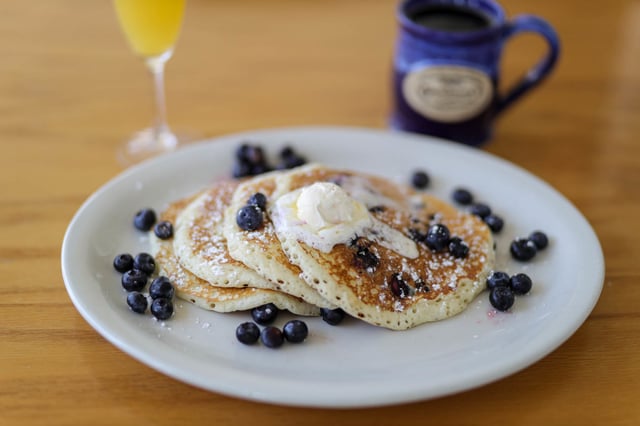 Stack of blueberry pancakes with butter and powdered sugar beside a mimosa and blue coffee mug
