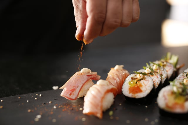 Chef's hand sprinkling spice over salmon and tuna nigiri beside sliced sushi rolls on dark slate