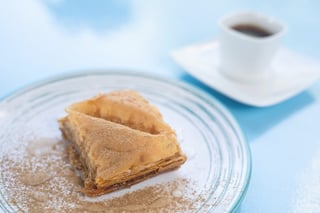 Slice of golden baklava dusted with cinnamon on a glass plate beside a cup of coffee