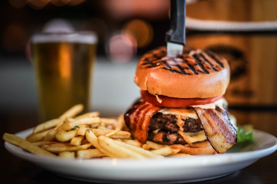 Grilled-bun bacon cheeseburger with a steak knife, a plate of fries and a golden beer behind