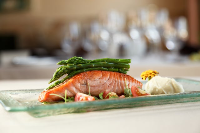 Seared salmon fillet topped with asparagus, tomatoes and purée on a glass plate before blurred glasses