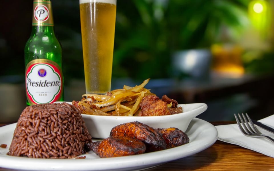Plate of black beans and rice, sweet plantains and fried pork with onions beside a cold Presidente beer