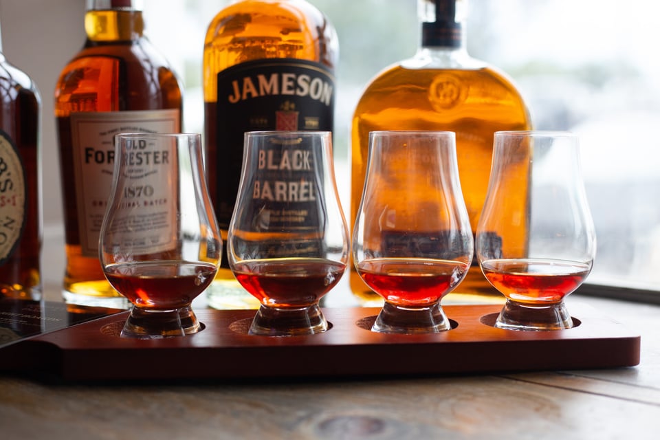 Flight of four whiskey tasting glasses on a wooden board before bottles of Jameson, Old Forester and Woodford Reserve