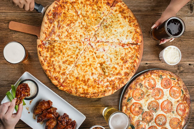 Overhead view of cheese and tomato pizzas, glazed wings and beers on a rustic wooden table
