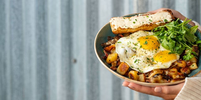 Hands holding a blue bowl of fried eggs, crispy potatoes, mushrooms, arugula and toast