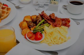 Plated breakfast of scrambled eggs, bacon, asparagus, potatoes and tomatoes beside coffee, orange juice and waffles