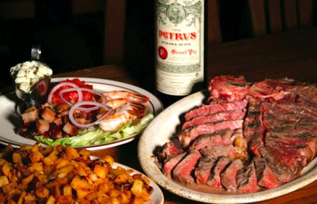 Platter of sliced rib steak beside shrimp salad, roasted potatoes and a bottle of Petrus wine