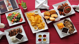 Overhead view of Cuban dishes including plantain chips, salad, grilled meats, rice and bread on a red tablecloth