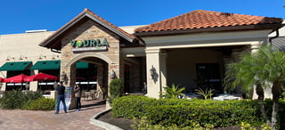 Sunlit Vourla Mediterranean Grill entrance with stone archway, terracotta roof, red umbrellas and covered patio tables
