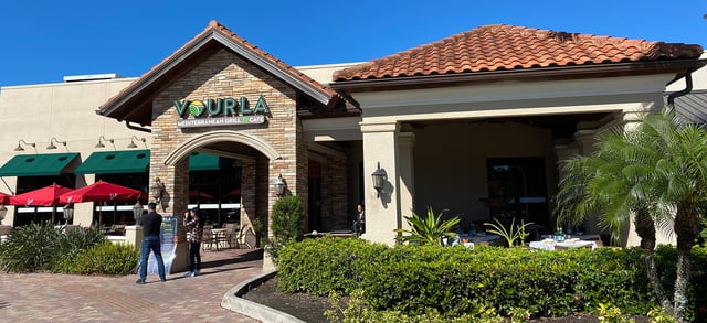 Sunlit Vourla Mediterranean Grill entrance with stone archway, terracotta roof, red umbrellas and covered patio tables