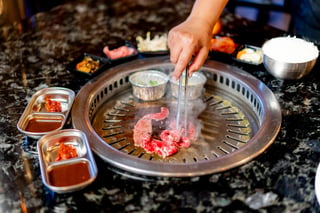 Hand using tongs to grill thin beef slices on a tabletop Korean barbecue surrounded by side dishes