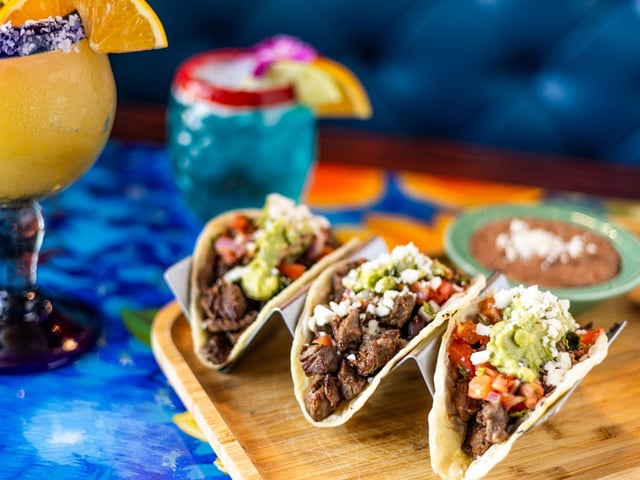 Three steak tacos topped with guacamole and cheese on a wooden board beside colorful margaritas