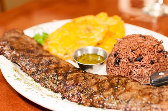 Grilled skirt steak with black beans and rice, fried plantains and green chimichurri sauce on a white plate