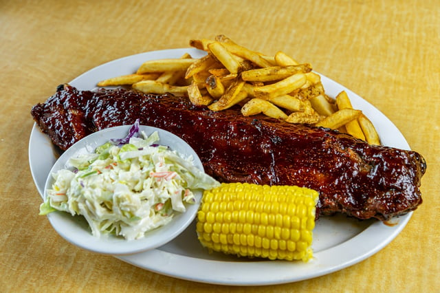 Full rack of barbecue-glazed ribs on an oval plate with fries, coleslaw and corn