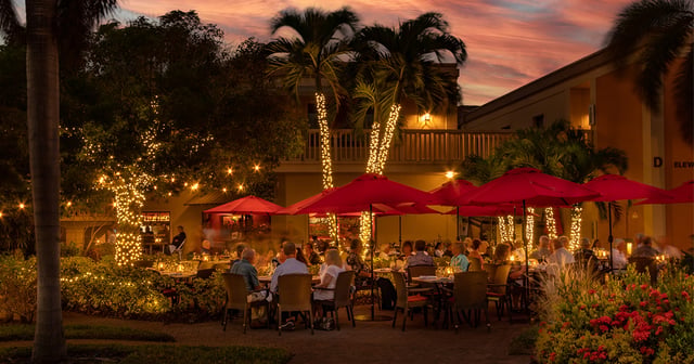 Diners seated under red umbrellas on a twilight garden patio with palm trees wrapped in string lights