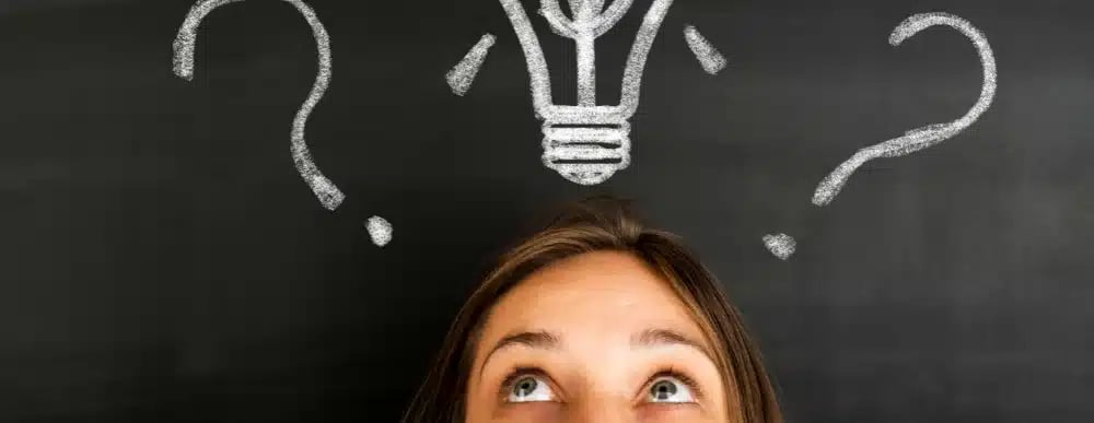 Person looking up at a light bulb and question marks drawn on a chalkboard,