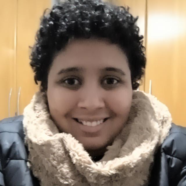 Foto de Louise Suelen sorridente de cabelos curtos e cacheados, vestindo casaco escuro e cachecol de lã bege, em ambiente interno com armários de madeira ao fundo.