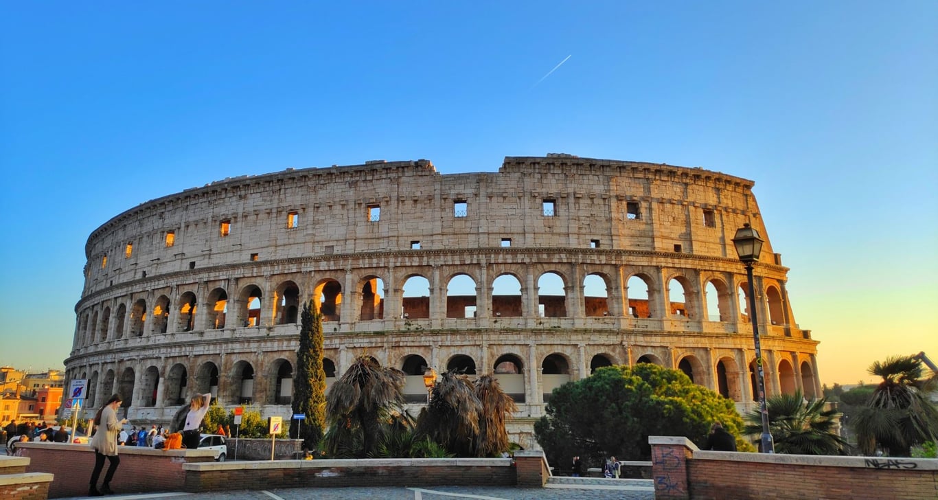 uma foto do Coliseu em Roma, Itália. O Coliseu é um grande anfiteatro oval com vários níveis de arcos. A foto foi tirada de longe, com o Coliseu ocupando a maior parte do quadro. O céu é azul e há palmeiras e outros prédios ao fundo. Existem algumas pessoas visíveis em primeiro plano, mas elas estão desfocadas e não são o foco da foto.
