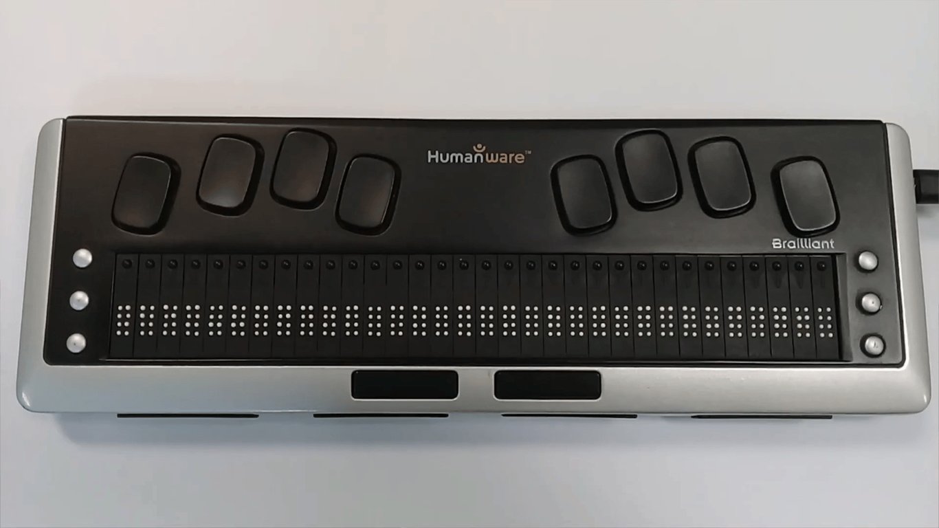 Linha Braille retangular, com acabamento prateado nas laterais e preto no centro. Ele possui uma fileira de 40 células Braille com pinos táteis e botões de entrada na parte superior, organizados em dois grupos de quatro para as mãos esquerda e direita. Dois botões retangulares alongados estão localizados na borda inferior.