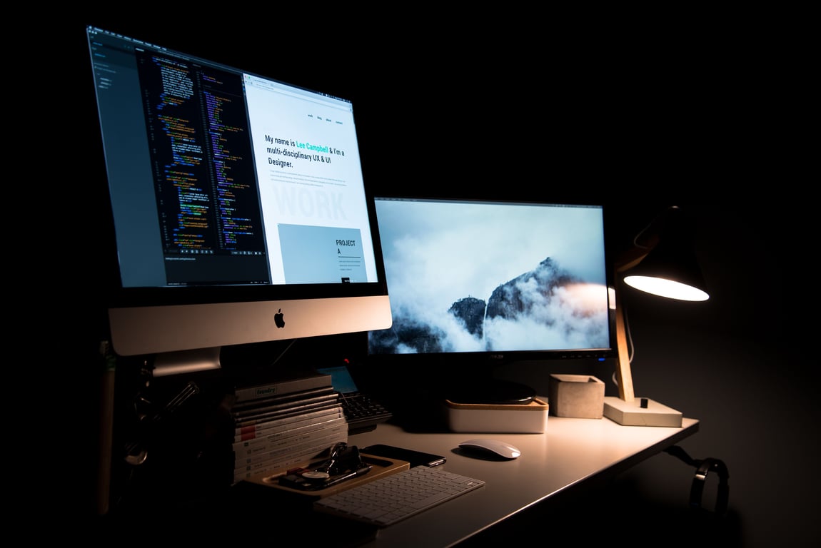 um ambiente de trabalho moderno e organizado. Há uma mesa com dois monitores. O monitor à esquerda exibe códigos de programação coloridos, enquanto o monitor à direita mostra uma paisagem serena de montanhas cobertas por nuvens. Uma lâmpada de mesa com design moderno ilumina a área de trabalho. Na frente dos monitores, há um teclado e um mouse brancos. Além disso, cadernos empilhados e outros itens pequenos estão organizados ao lado do teclado.