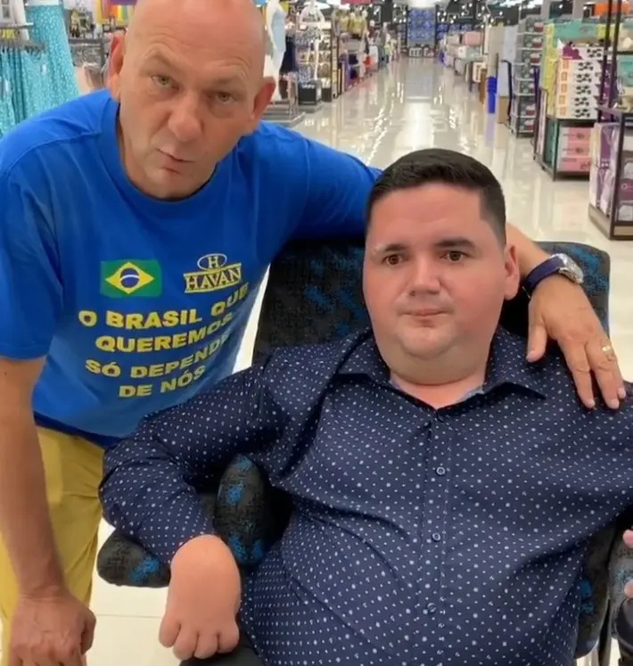 Luciano Hang, empresário e dono da rede de lojas Havan, aparece em pé ao lado de Jelres Freitas, advogado e então presidente da Comissão dos Direitos das Pessoas com Deficiência da OAB de Sumaré (SP), que está sentado em uma cadeira de rodas. A foto é tirada dentro de uma loja Havan, bem iluminada, com prateleiras e corredores ao fundo. Luciano Hang veste uma camiseta azul com mensagens patrióticas e o logotipo da Havan, enquanto Jelres Freitas usa uma camisa social azul-marinho com estampa discreta.