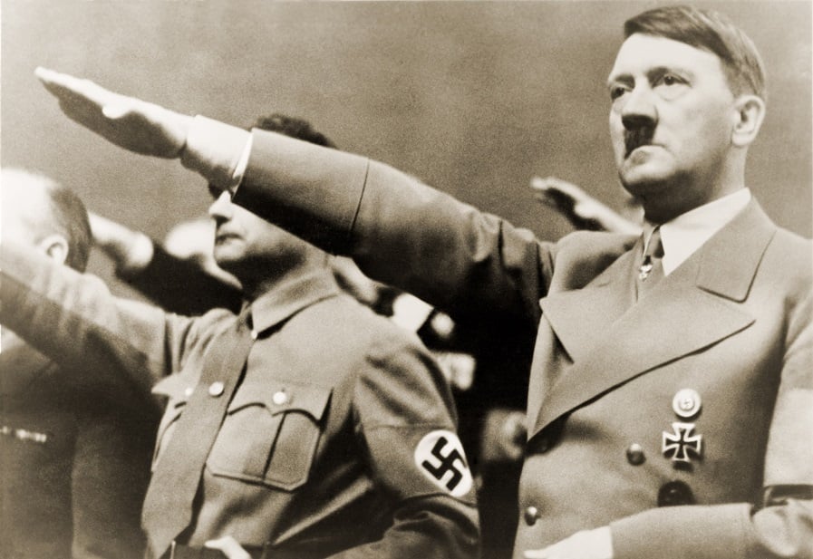 Fotografia histórica em preto e branco mostra Adolf Hitler em primeiro plano, de uniforme nazista, com expressão séria e braço direito estendido para frente, realizando a saudação nazista. Atrás dele, outros homens também fazem o mesmo gesto. Na manga de um deles aparece uma braçadeira com a suástica, símbolo do regime nazista. A imagem representa um momento de propaganda do Terceiro Reich.