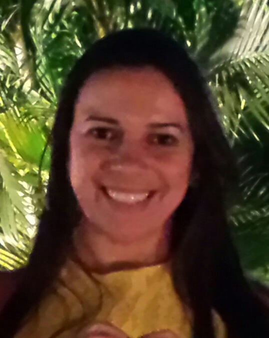Foto da Professora Ana Célia sorridente de cabelos longos e escuros, usando uma roupa amarela, em frente a folhagens verdes iluminadas.