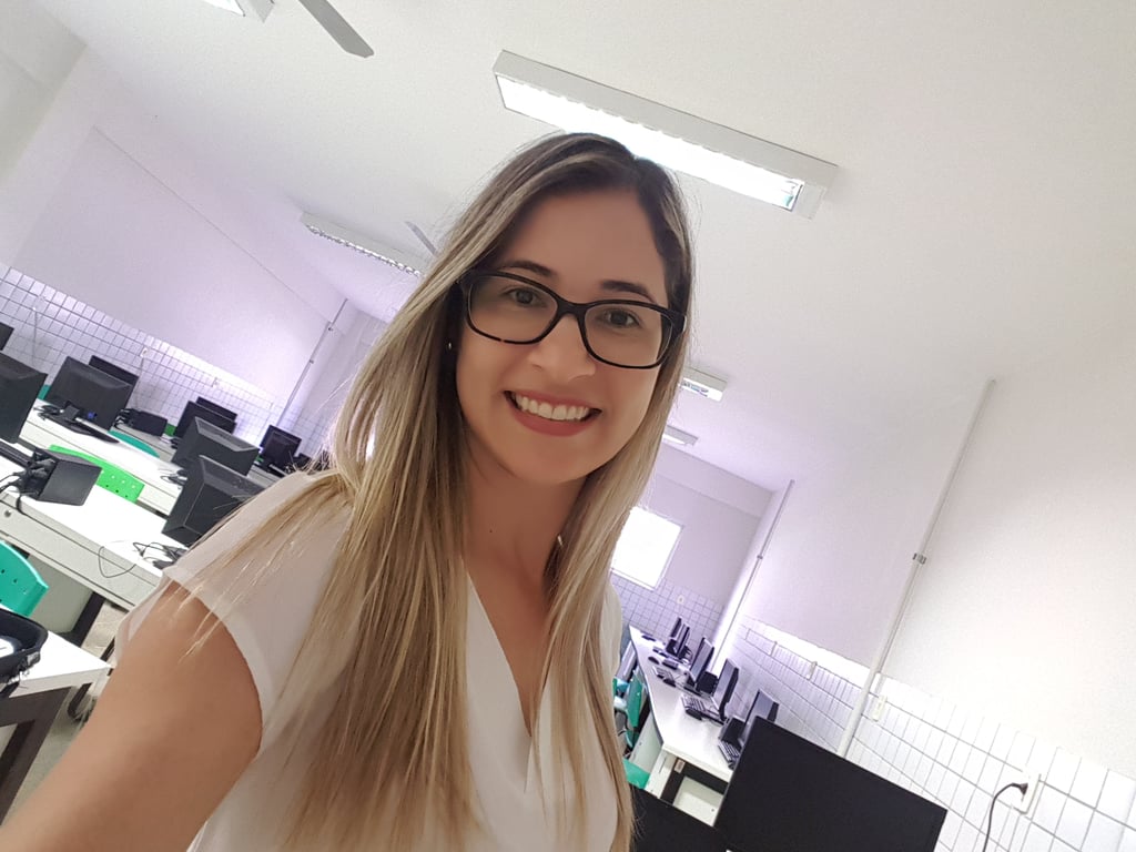 Foto da Professora Karine sorridente de cabelos longos e loiros, usando óculos e blusa clara, em uma sala de informática com vários computadores dispostos em mesas.